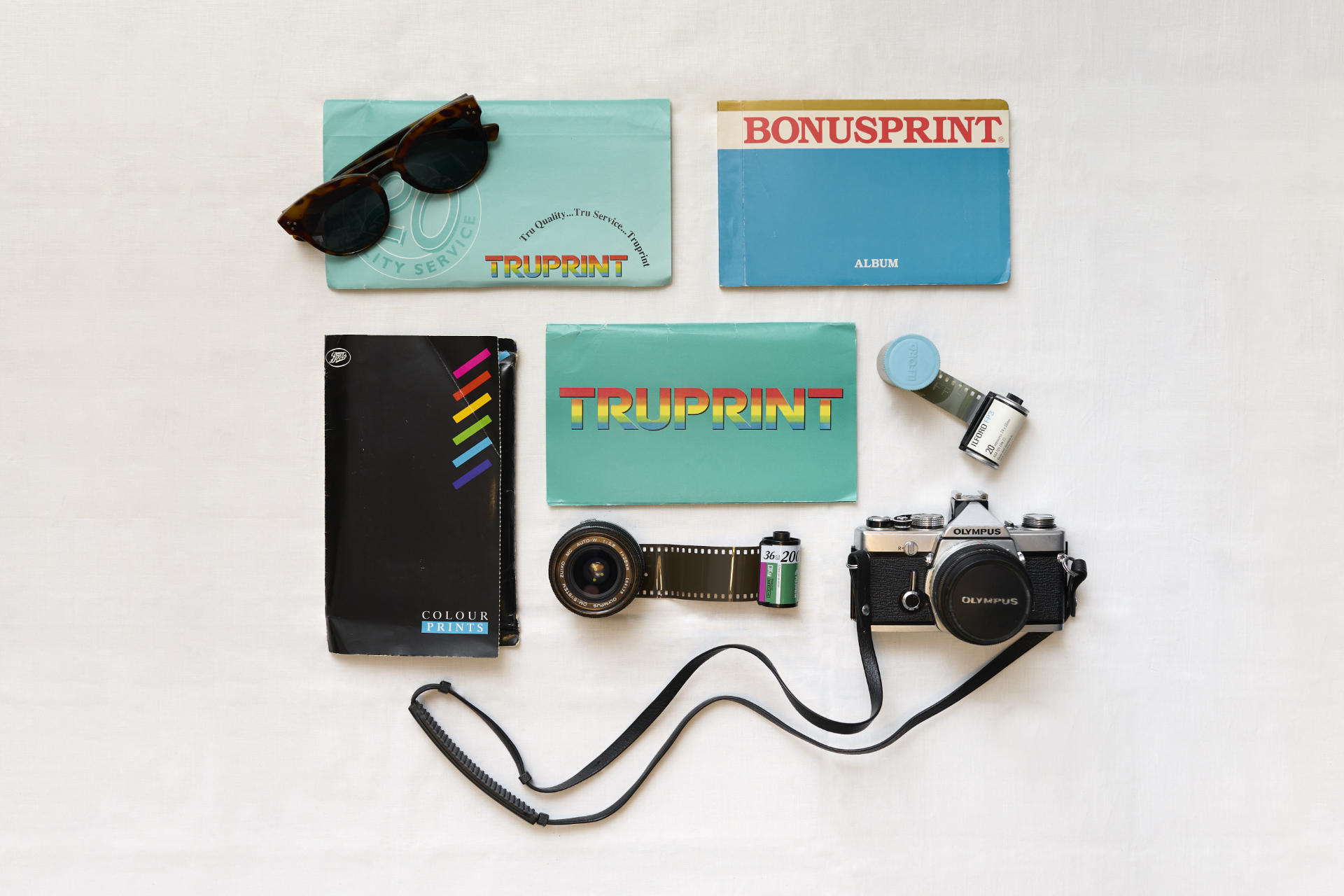 A flatlay of old 1990s 6x4 inch photograph wallets from high-street photo labs and chemists, a pair of sunglasses, some old 35mm film reels, canisters and pots, a vintage Olympus Zuiko camera lens, and an Olympus OM-1N film camera on a white background